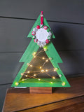Green-painted pine Christmas tree with walnut base, glowing fairy lights and hanging holiday photo-frame ornament