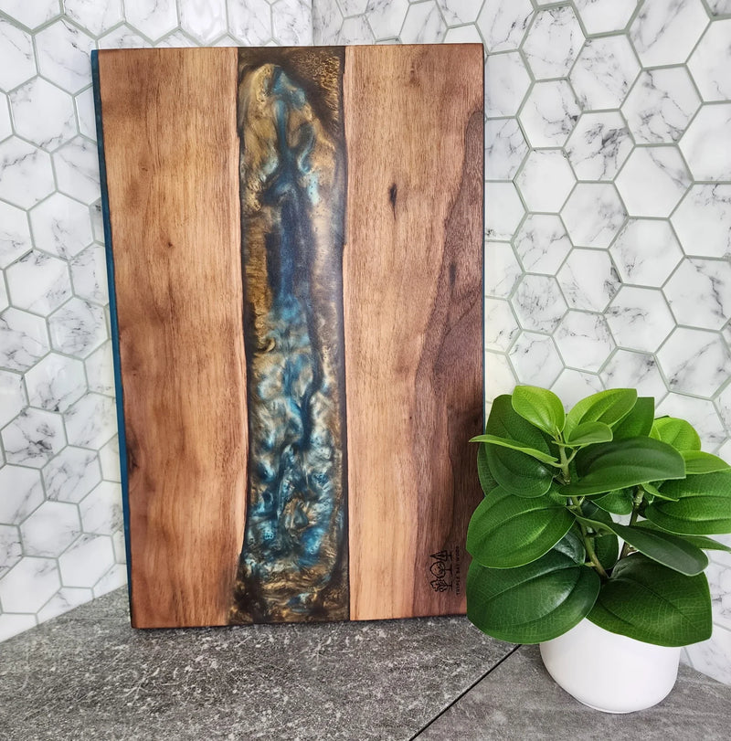 Handcrafted wood and resin cutting board with blue and gold accents, styled on a kitchen counter with a potted plant.