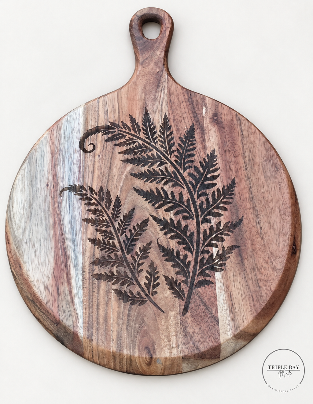 Round acacia serving board with dark fern frond engraving, pale wood bands and oval hole in the paddle handle