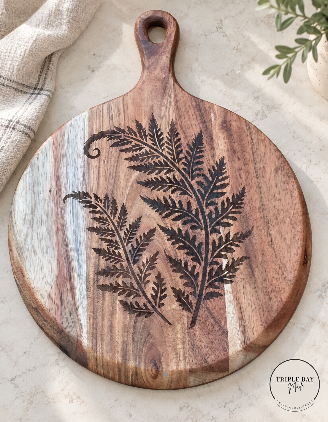 Fern-engraved round acacia serving board styled on light stone beside neutral linen and greenery