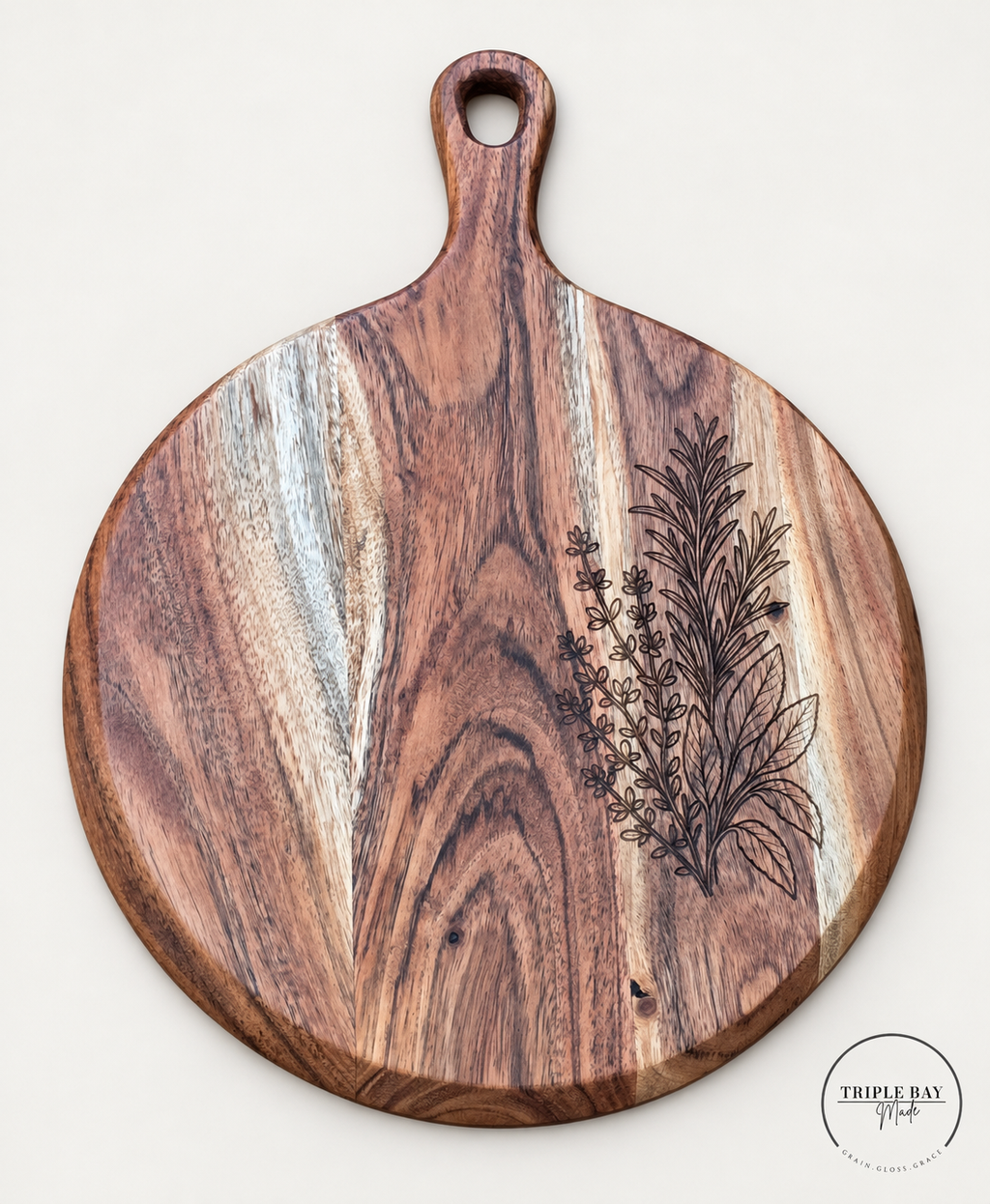 Round acacia serving board with fine herb sprig engraving along the right side and an oval hanging hole