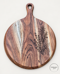 Round acacia serving board with fine herb sprig engraving along the right side and an oval hanging hole