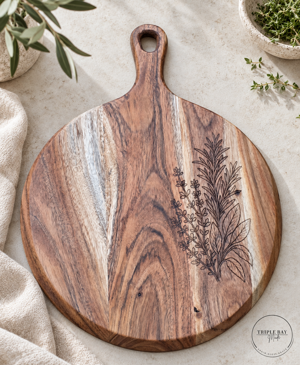 Herb-engraved acacia serving board on light stone beside cream linen and potted greenery