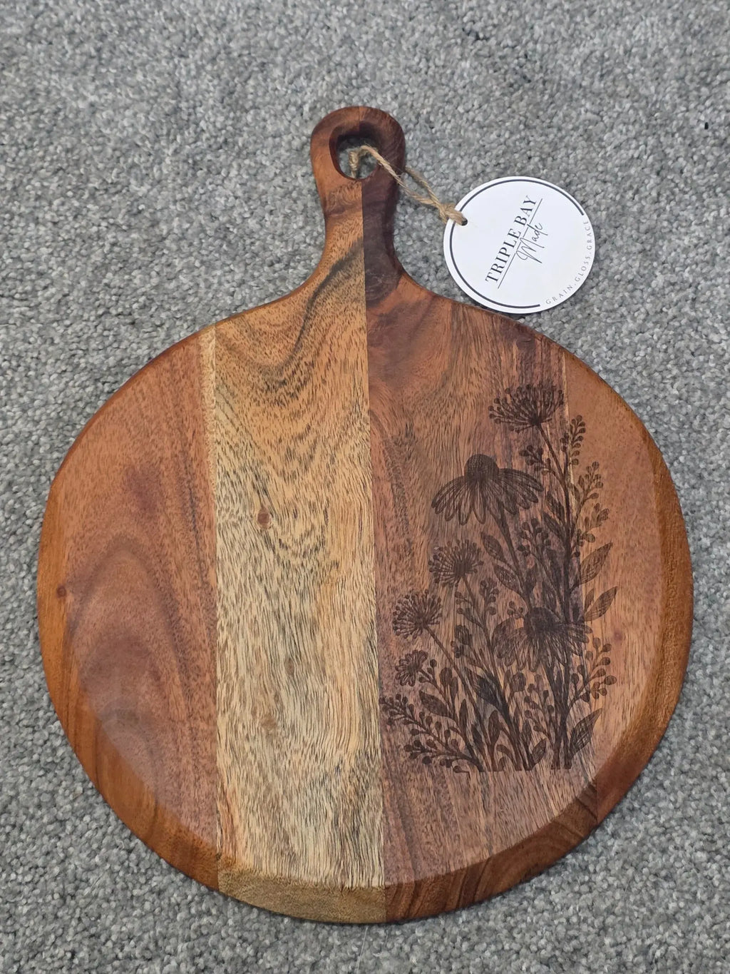 Round acacia paddle serving board with wildflowers and leafy stems engraved along the right side