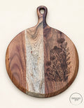 Round acacia serving board with wildflower engraving along the right edge, pale central grain band and paddle handle