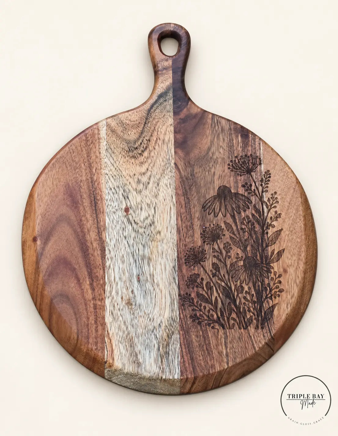 Round acacia serving board with wildflower engraving along the right edge, pale central grain band and paddle handle