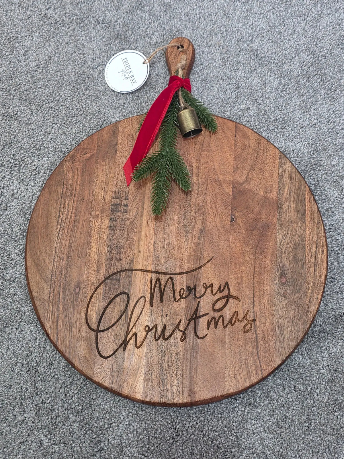 Round acacia board with Merry Christmas script engraving, handle, red ribbon, greenery and bell
