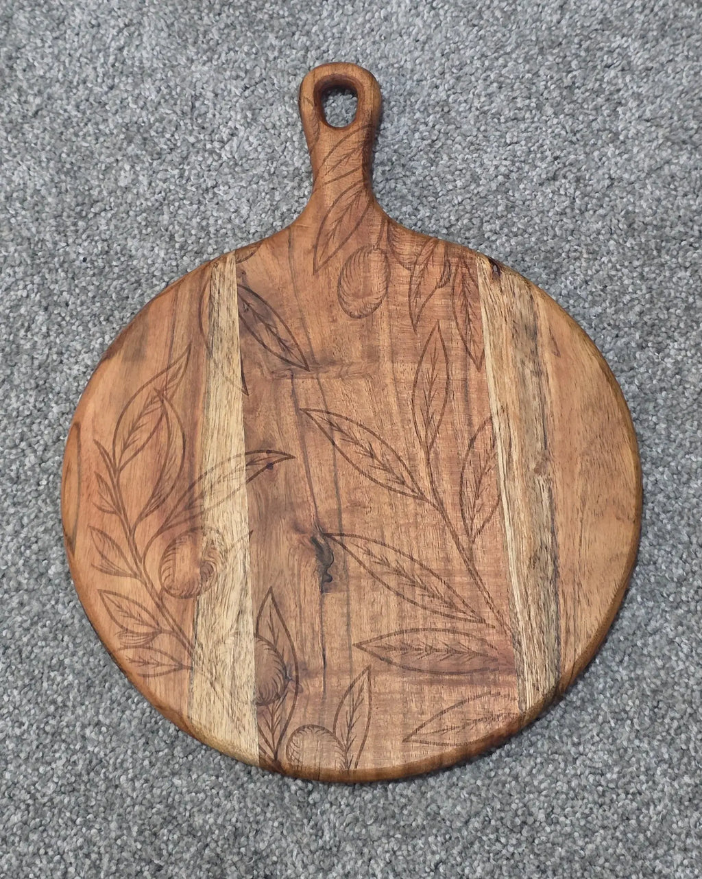 Round acacia paddle serving board with fine leaf and hanging fruit engraving across contrasting wood grain