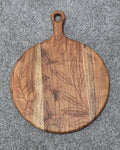 Round acacia paddle serving board with fine leaf and hanging fruit engraving across contrasting wood grain