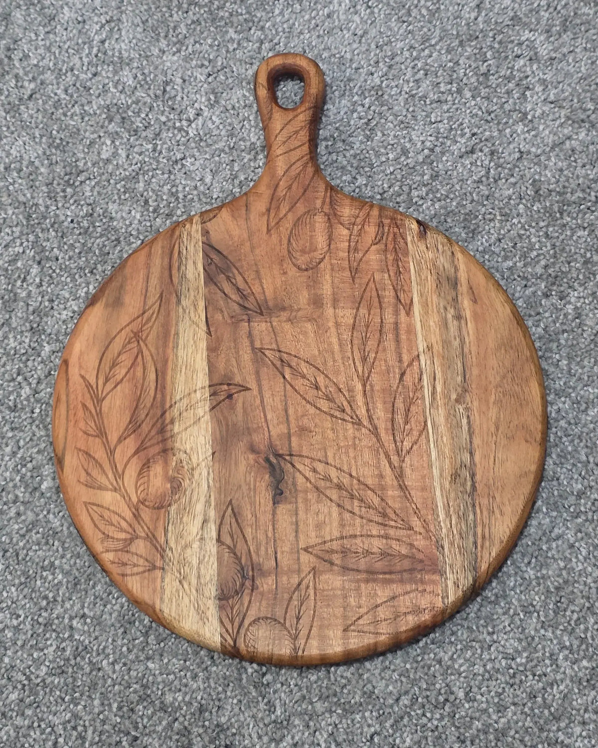 Round acacia paddle serving board with fine leaf and hanging fruit engraving across contrasting wood grain