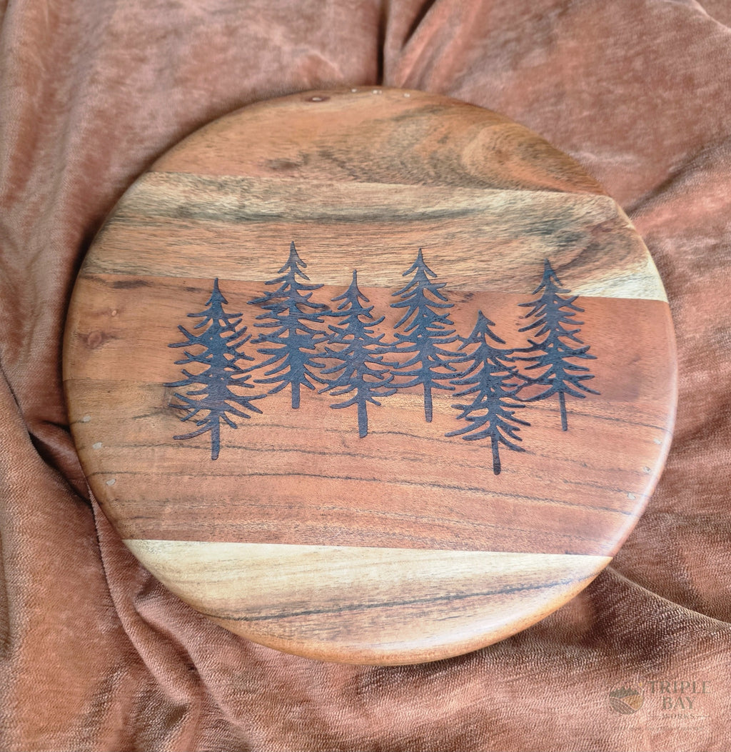 Round acacia footed serving board engraved with a grove of evergreen trees