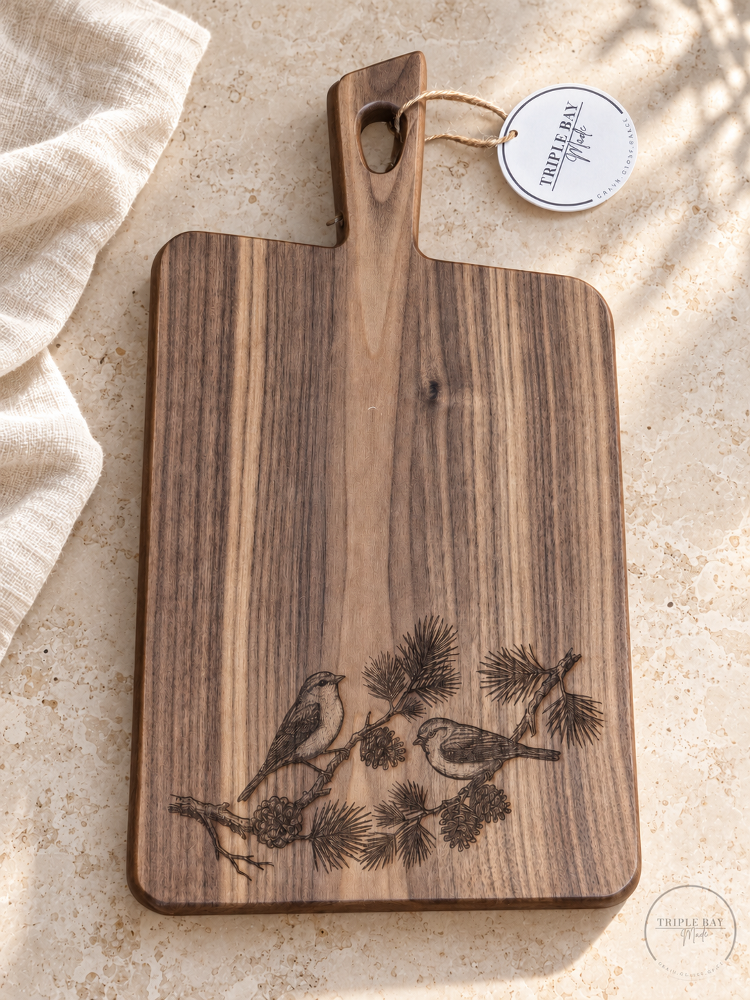 Bird and pine engraved acacia board with walnut finish styled on light stone beside ivory linen