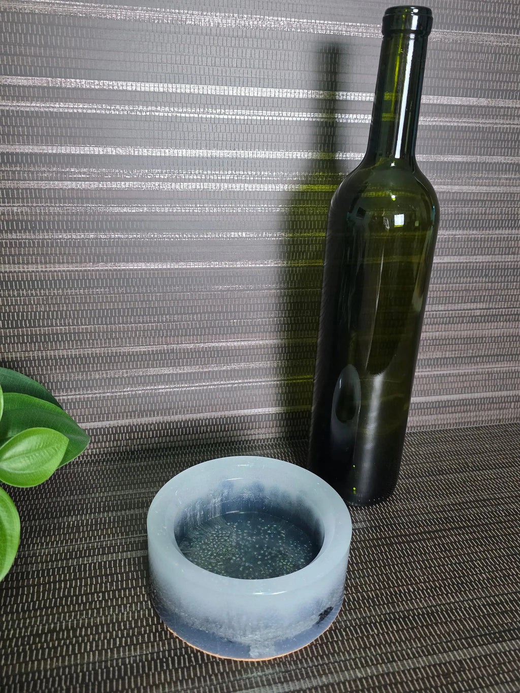 Green glass bottle and white ceramic object on a textured surface with a plant in the corner.