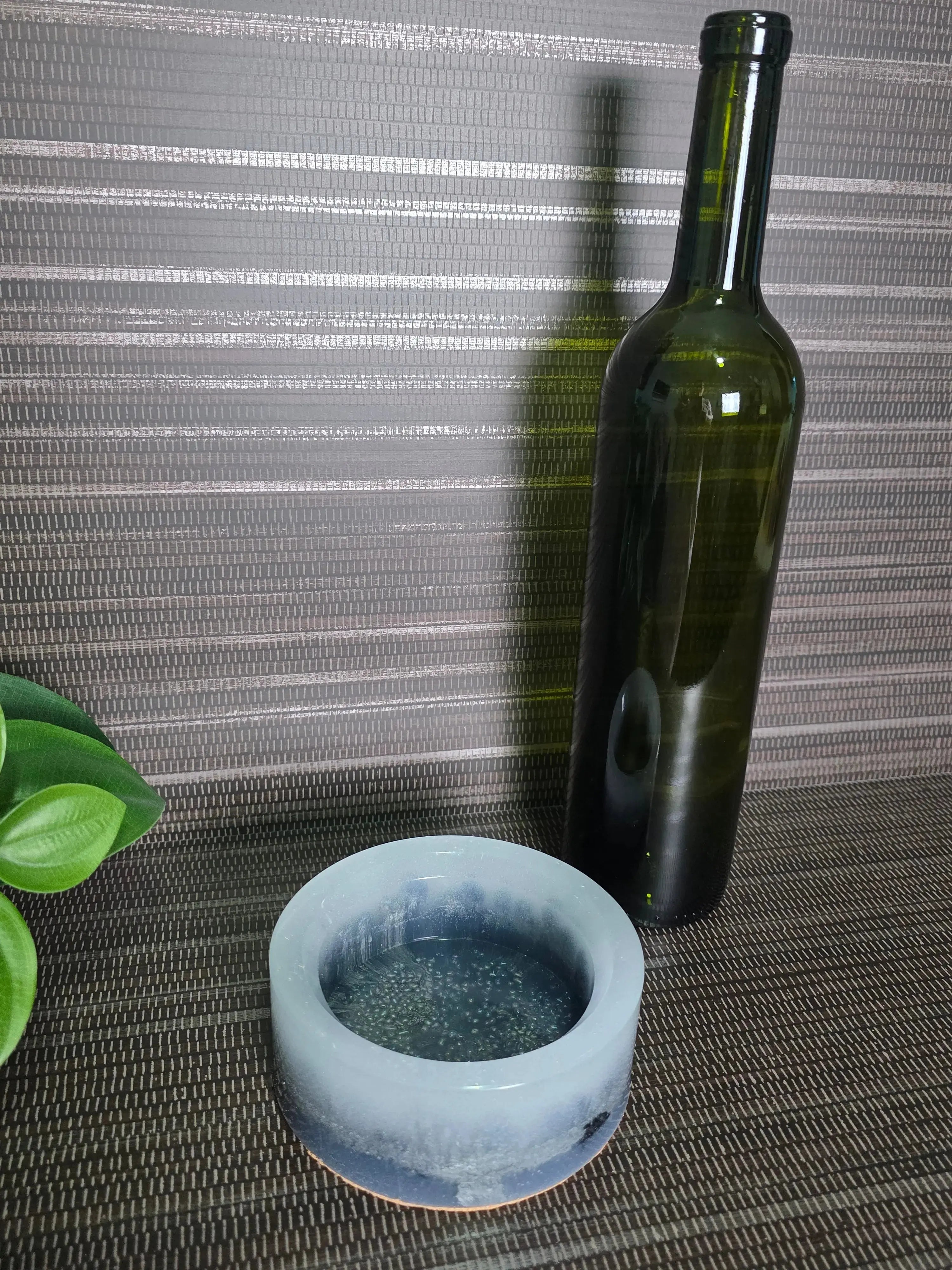 Green glass bottle and white ceramic object on a textured surface with a plant in the corner.