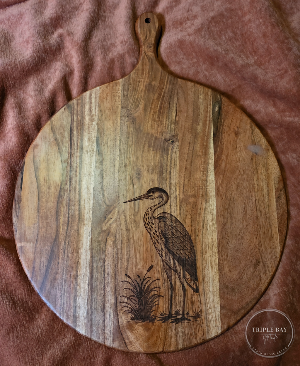 Round acacia paddle serving board with a heron and cattail engraving on the front