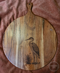 Round acacia paddle serving board with a heron and cattail engraving on the front