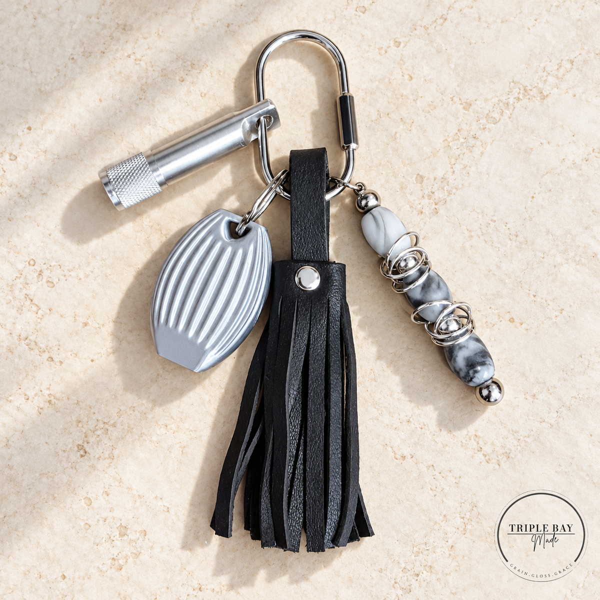 Black real leather tassel charm laid on ivory stone, showing the silver-tone clip, keychain light, dual-tone pebble whistle, and three gray-and-white beads with fidget rings.