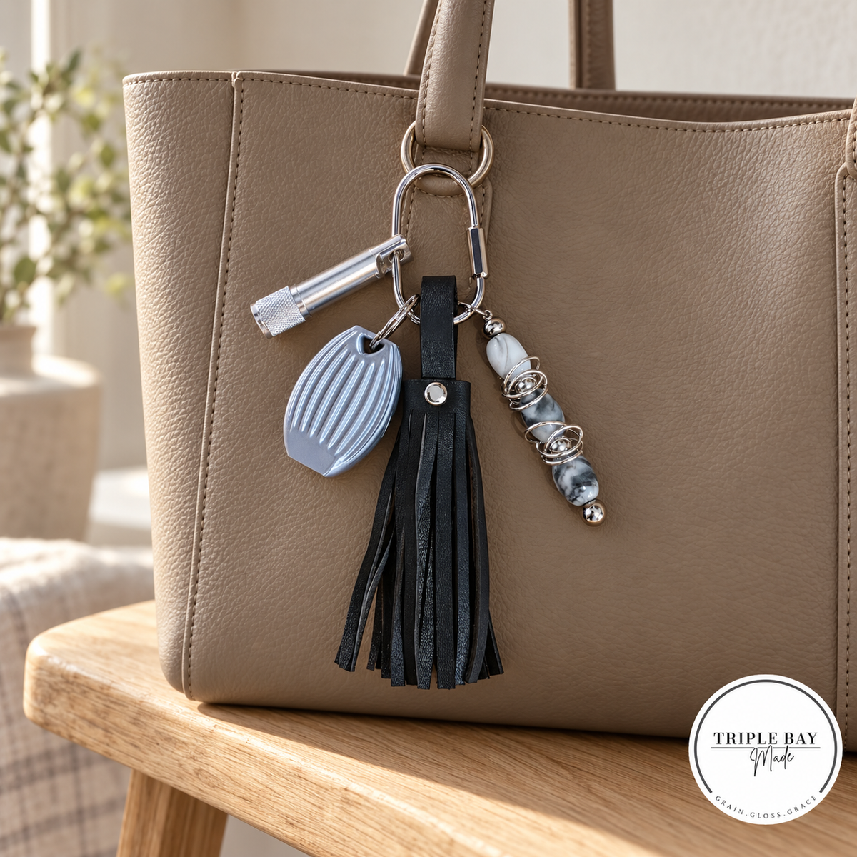 Seven-inch black leather tassel bag charm clipped to a taupe tote, with a silver-tone keychain light, pebble whistle, and gray-and-white fidget beads; tote is a styling prop.